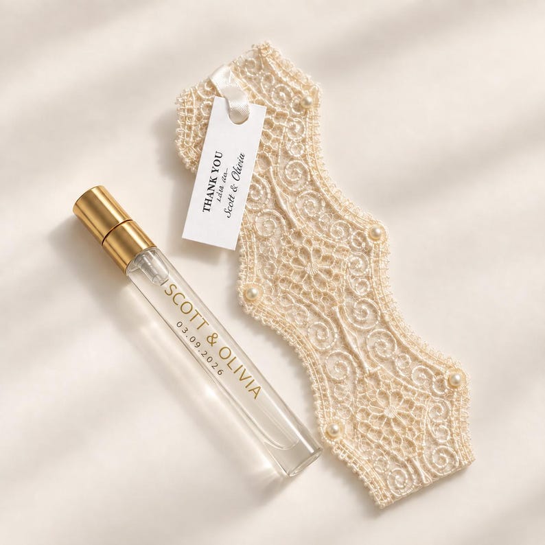A clear glass perfume atomiser with a gold cap, featuring the text SCOTT & OLIVIA and 03.09.2026. Attached is a white tag reading THANK YOU with Scott & Olivia, alongside a cream-coloured lace fabric piece adorned with small white pearls.