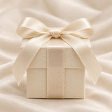 A square, cream-coloured gift box with a textured surface, wrapped in a matching satin ribbon tied into a bow on top. The box features a clean, structured design with a lid that sits flush against the base, centred with a smooth ribbon.